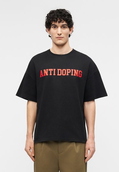 Jeune homme aux cheveux bouclés et foncés portant un T-shirt noir avec l'inscription « ANTI DOPING » en lettres rouges, associé à un pantalon vert olive.