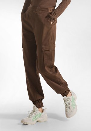 Bruine cargobroek met taps toelopende enkels, zijzakken en een duurzame stof. Gepaard met lichte sneakers met groene accenten.