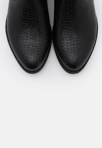 Un par de zapatos de cuero negro con un patrón de piel de cocodrilo, vistos desde arriba sobre un fondo blanco.