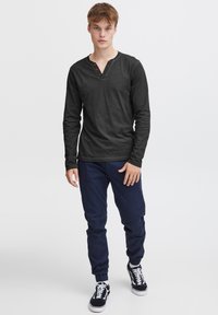 Langærmet sort henley-skjorte med V-hals og tre brune knapper. Sammen med marineblå bukser og sorte sneakers. Enkel design.