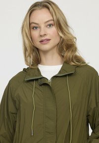 Veste légère vert olive avec fermeture éclair sur le devant, capuche ajustable et cordons de serrage. Texture lisse et accents de couture visibles.