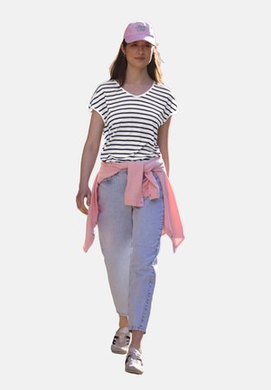 Ung kvinde går, iført en hvid og sort stribet skjorte, lyseblå jeans, lyserød sweater bundet om livet, lyserød kasket og sneakers.