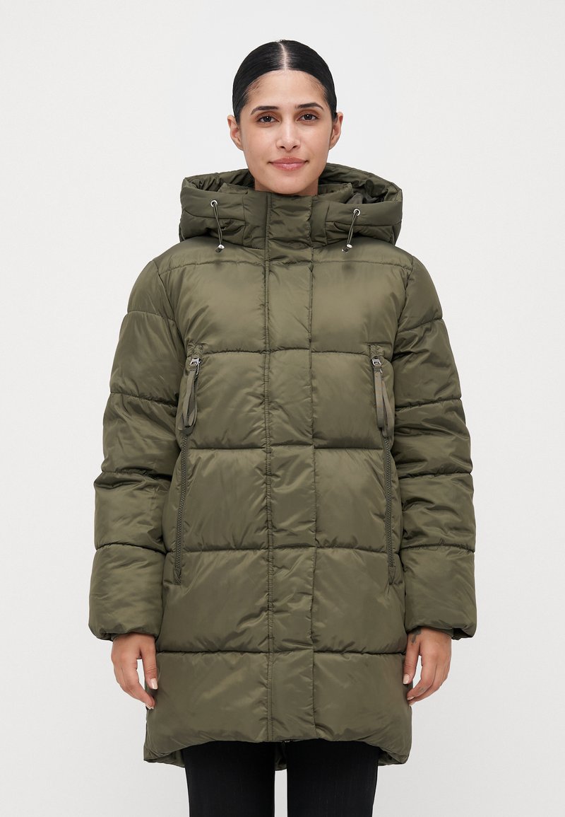 Mujer vestida con una chaqueta acolchada en color verde oliva, con capucha y bolsillos con cremallera, de pie contra un fondo claro y liso.