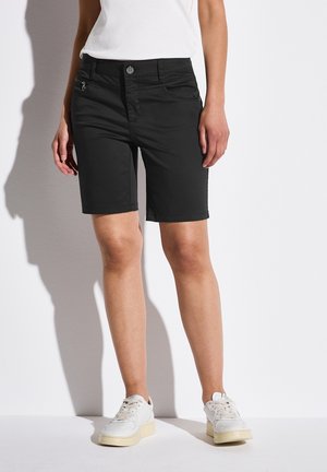 Person iført sorte knælange shorts, en hvid top og hvide sneakers, der står mod en lys baggrund.