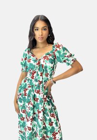 Robe maxi florale en vert, rouge et blanc ; manches courtes bouffantes ; corsage ajusté avec un détail noué ; jupe fluide ; tissu en coton.