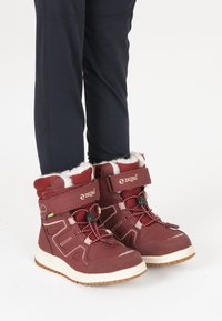 Les bottes d'hiver bourgogne présentent un matériau synthétique avec une doublure en fourrure blanche, des accents noirs et blancs, un système de laçage et une semelle en caoutchouc.