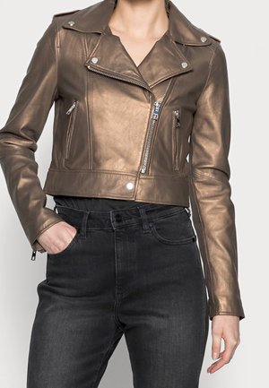 Korte bronzen leren bikerjas met asymmetrische ritssluiting, ritssluitingszakken op de borst, gedragen met zwarte hoge taille jeans, hand in de zak.