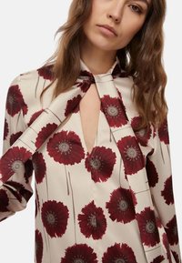 Blusa bianca con motivo floreale rosso, presenta un collo a keyhole e un dettaglio con laccetto. Realizzata in tessuto liscio con una leggera lucentezza.