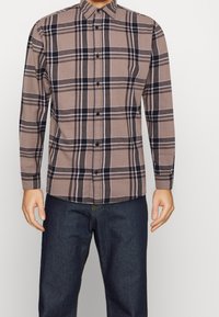 Mężczyzna w brązowej koszuli w kratę z długim rękawem, zapinanej na guziki, w połączeniu z ciemnoniebieskimi dżinsami, stojący na jednolitym tle.