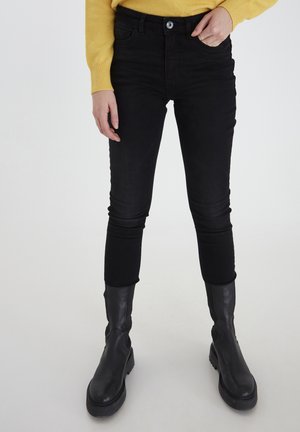 Person trägt schwarze Skinny Jeans, schwarze Stiefel bis zur Mitte der Wade und einen gelben Pullover, steht vor einem schlichten hellen Hintergrund.