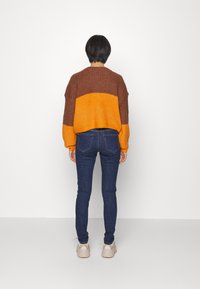 Stickad tröja med en brun övre del och en orange nedre del, tillsammans med mörka denimjeans och beige sneakers. Tätt passform.