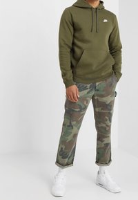 Oliivinvihreä huppari, jossa etutasku ja valkoinen logo, yhdistettynä maastokuvioisiin housuihin sekä valkoisiin tennareihin ja ribbineulottuihin nilkkasukkiin.