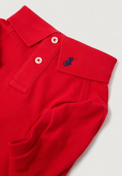 Rdeča polo majica z teksturirano površino, tri gumbi na plastronu in modrim vezenim logotipom na ovratniku. Ima kontrastne gumbe.