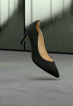 Zapato de tacón alto negro con punta puntiaguda, borde festoneado y interior marrón claro flotando sobre suelo gris con fondo verde desenfocado.