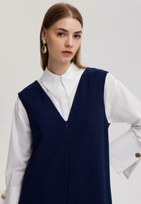 Jūras zils bezpiedurkņu kleita ar V veida kakla izgriezumu, vilkta virs baltas apkakles krekla. Tam ir priekšējais rāvējslēdzējs un zelta pogu akcenti uz krekla aprocēm.