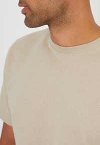 Beige texturované tričko s kulatým výstřihem a krátkými rukávy, s jemnými horizontálními pruhy a žebrovaným detailem u límce.