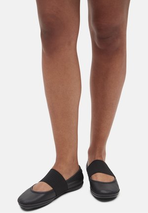 Black flat shoes with a soft leather upper, open toe design, and wide elastic band across the instep, resting on a flexible sole.