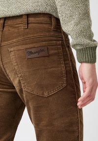 Person som bär bruna kordflors Wrangler-byxor och en stickad tröja med gröna manschetter, visar bakfickan och handflatan.