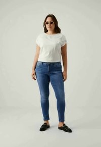 Top blanco de manga corta, jeans ajustados azules, mocasines negros. La modelo se encuentra de pie con los brazos relajados en un fondo neutro. Texturas suaves, diseño simple.