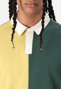 Camisa polo con secciones amarillas y verdes, con un cuello blanco en contraste. Material de algodón, superficie texturizada, con un diseño de tapeta central.