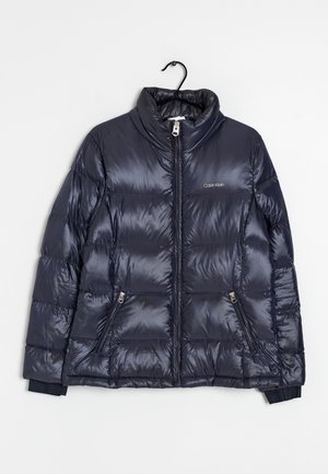 Marineblå Calvin Klein dunjakke med høj krave og lynlåslommer i siderne hængende på en sort bøjle.