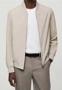 Beige Bomberjacke mit einfarbigem Design, gerippten Bündchen und einem Reißverschluss vorne. Getragen über einem weißen T-Shirt und beigen Hosen, wodurch eine neutrale Farbpalette betont wird.