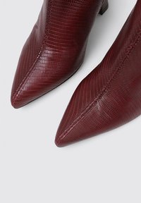 Botines de tobillo burdeos con punta afilada, fabricados en cuero sintético texturizado, que presentan un diseño elegante con un bajo tacón cuadrado y detalles de costura sutiles.