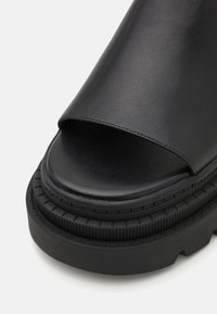 Sandale slide noire à bout ouvert avec une semelle épaisse texturée et une bride supérieure lisse couvrant l'avant du pied.