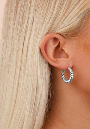 Silverfärgade hoopörhängen med flätad textur, utrustade med en klämlås. Visas på en models öra med rakt, blont hår.