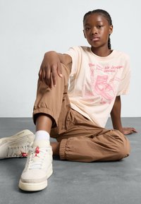 Camiseta beige clara con estampado gráfico rosa, combinada con pantalones marrones de puños elásticos y zapatillas color crema con acentos rojos.