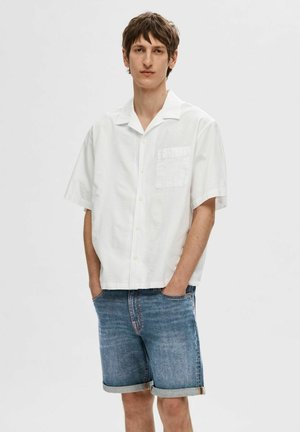 Hvid kortærmet skjorte med knapper og lomme, parret med blå denimshorts rullet ved kanten. Enkel, afslappet pasform.