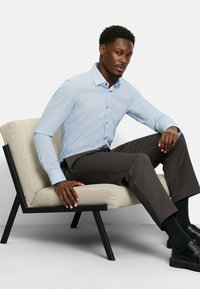 Mann in hellblauem, gemustertem Hemd und dunkler Hose sitzt auf einem beigen gepolsterten Stuhl mit schwarzen Beinen vor einem schlichten Hintergrund.