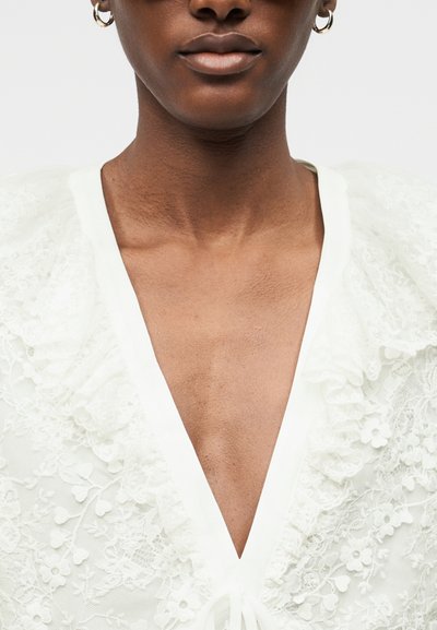 Gros plan sur une personne portant un vêtement en dentelle blanche avec des motifs floraux et un décolleté en V profond, accessoirisé avec des boucles d'oreilles créoles dorées.