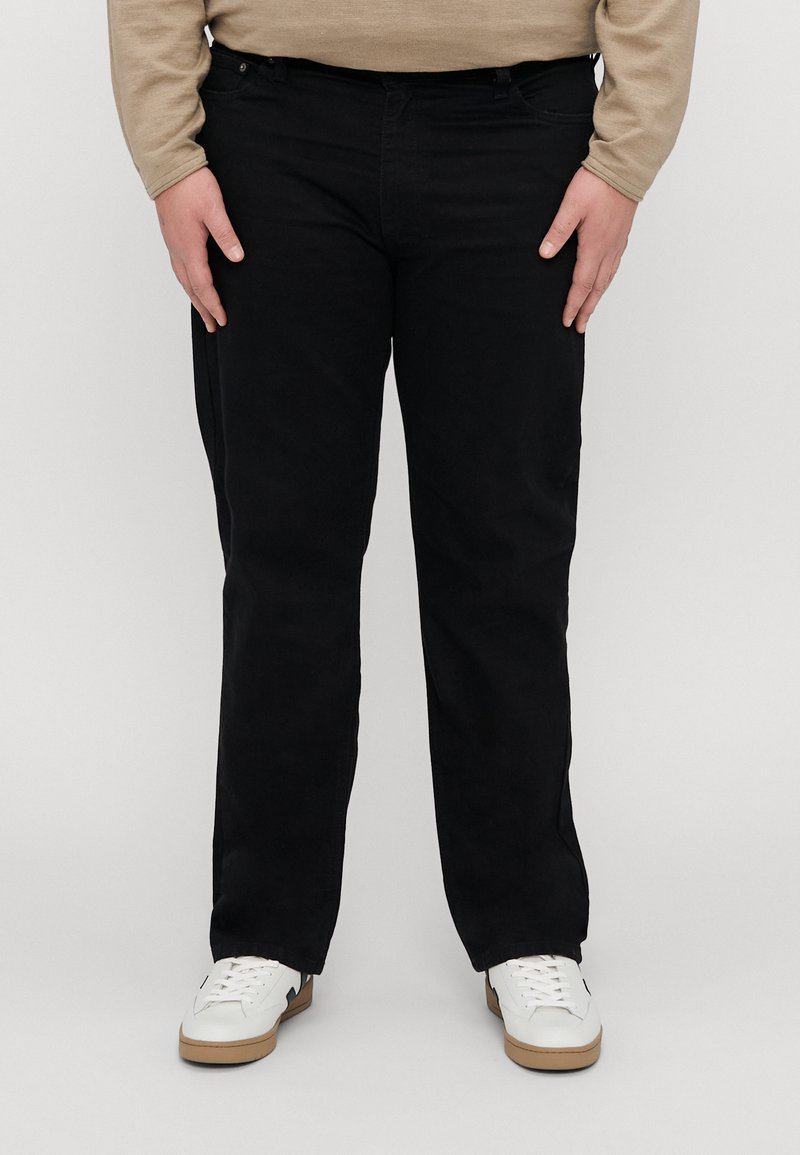Sorte bomullsbukser med rett ben-design, som har sidelommer og beltehemper, kombinert med hvite joggesko mot en grå bakgrunn.