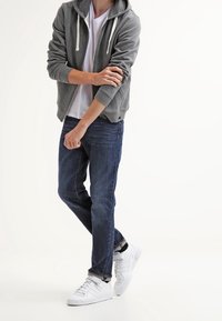 Grå zip-up hoodie med dragsko, vit t-shirt under, blå denim jeans och vita sneakers med svarta detaljer.