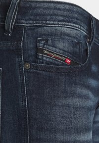 Mörkblå denimjeans i närbild som visar myntfickan med Diesel Industry-märket och nitar.