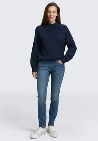 Marineblauwe geribbelde trui met hoge hals en verlaagde schouders, gecombineerd met strakke blauwe jeans en witte sneakers. Eenvoudige, casual outfit.