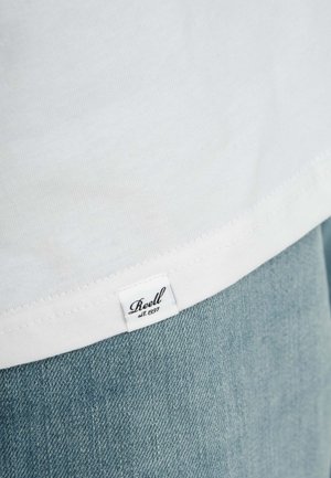 Tiro de una camiseta de algodón blanca con el dobladillo visible y una textura de tela, junto a una etiqueta que detalla "Reell est. 1997", combinada con jeans de mezclilla azul claro.