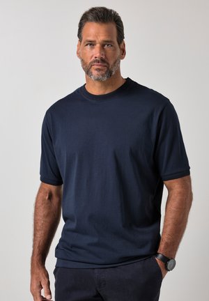 Uomo di mezza età con barba corta che indossa una maglietta blu navy e pantaloni scuri, in piedi su uno sfondo chiaro e semplice.