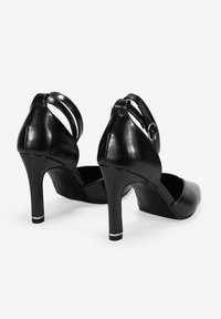 Zapatos de tacón alto negros con punta redondeada y correa en el tobillo. Fabricados en cuero brillante con una textura suave y un acento plateado en el tacón.