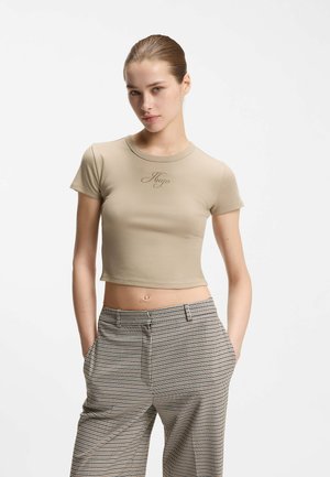 Woman with slicked-back hair wears a beige cropped T-shirt with "Hugo" text and high-waisted checkered trousers, hands in pockets, neutral background.