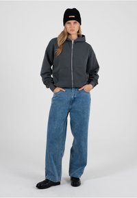 Grauer Kapuzenpullover mit Reißverschluss und Fronttaschen, kombiniert mit hoch taillierter blauer Jeans und schwarzen Schuhen. Eine schwarze Strickmütze vervollständigt den Look.