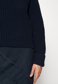 Granatowy dziergany sweter o prążkowanej fakturze i szerokich mankietach, zestawiony z ciemną, falbaną spódnicą. Skupienie na tkanym wzorze tkaniny.