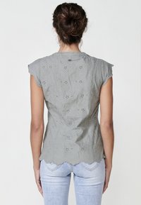 Hellgraue Bluse mit floralem Stickmuster, gewellten Kanten und kurzen Puffärmeln. Der Stoff wirkt weich und hat eine strukturierte Oberfläche.