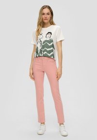 Camiseta blanca con un diseño gráfico en negro y verde de dos figuras, combinada con unos vaqueros ajustados rosa claro y zapatillas blancas.