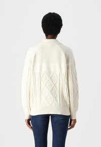 Weißer gestrickter Pullover mit einer Mischung aus Ripp- und Zopfmuster, in leicht oversized Passform und mit Kragen, kombiniert mit dunklen Jeans.