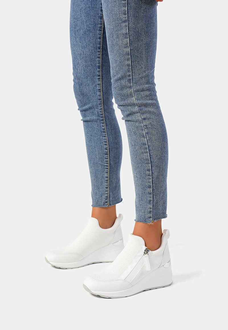 Sneaker bianche senza lacci con una tomaia texturizzata, dotate di una cerniera laterale e una suola a zeppa. Indossate con jeans aderenti blu.