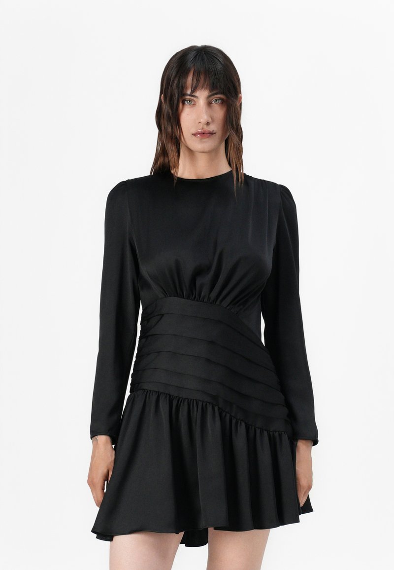 Schwarzes Kleid aus Satin mit figurbetontem Oberteil, gerafftem Taillenbund und Rüschensaum. Lange Ärmel und horizontale Falten an der Taille.