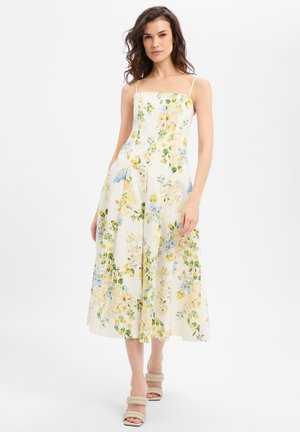 Frau trägt ein weißes Midikleid mit gelbem und blauem Blumenmuster, dünnen Trägern und beigen offenen Schuhen, steht vor einem weißen Hintergrund.