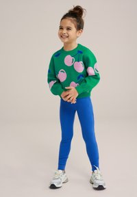 Zelený sveter s ružovými višňovými motívmi, v kombnácii s modrými legínami. Tento outfit má uvoľnený strih a rebrované manžety, doplnený o tenisky.